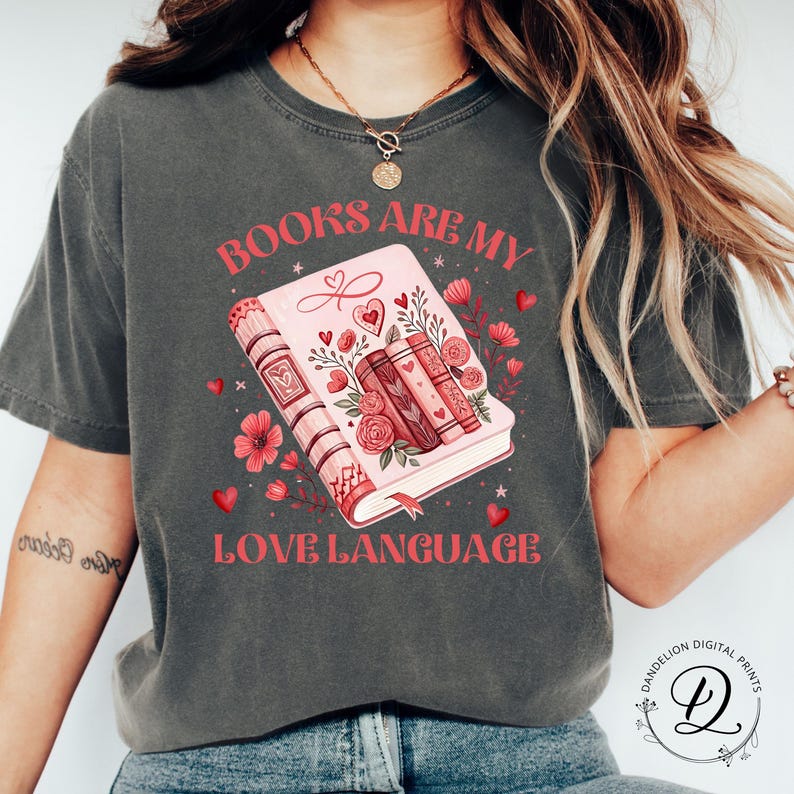 May include: Dark grey t-shirt with the text "BOOKS ARE MY LOVE LANGUAGE" in pink. The graphic features a pink book with floral and heart accents. The design includes a stack of books and decorative flowers.