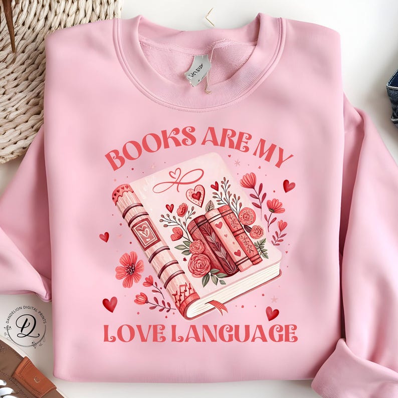 May include: Pale pink sweatshirt with the slogan "BOOKS ARE MY LOVE LANGUAGE" in red. The design includes a stack of books decorated with flowers and hearts. Ideal for book enthusiasts.