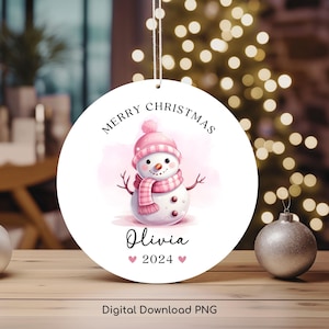 Könnte beinhalten: Ein runder weißer Weihnachtsbaumschmuck mit einem rosa Schneemann, der einen rosa Hut und einen Schal trägt. Der Schmuck trägt die Aufschrift "Merry Christmas" und "Olivia 2024" in schwarzer Schrift.