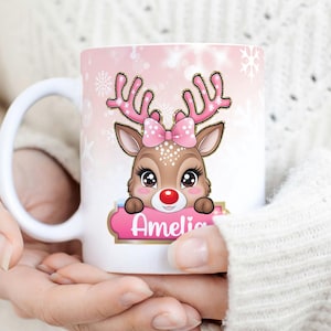 May include: White ceramic mug with a pink and gold reindeer design. The reindeer has a pink bow and a red nose. The mug says "Amelia" on a pink banner.