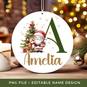 Op de afbeelding: Een ronde witte kerstornament met een grote donkergroene letter A, een cartoon kerstman, een klein rendier, een versierde kerstboom en de naam Amelia in gouden letters, opgehangen aan een wit koord tegen een wazige achtergrond.