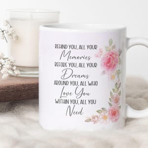Peut inclure: Un mug en c&eacute;ramique blanche orn&eacute; d'un motif floral de roses roses et de feuilles vertes sur le c&ocirc;t&eacute; droit. Le texte indique : Behind you, all your memories, before you, all your dreams, around you, all who love you, within you, all you need.