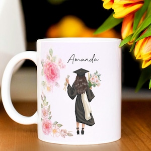Peut inclure: Un mug en c&eacute;ramique blanche orn&eacute; d'une illustration imprim&eacute;e repr&eacute;sentant une personne en toge et toque de dipl&ocirc;m&eacute; noire, tenant un dipl&ocirc;me et des fleurs. Le pr&eacute;nom Amanda est inscrit au-dessus, entour&eacute; de motifs floraux roses et d'un papillon.