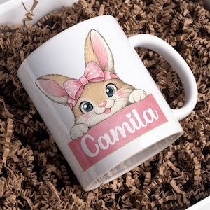 Puede incluir: Taza de cerámica blanca con un conejito de dibujos animados con un lazo rosa y el nombre "Camila" en una pancarta rosa. El conejito tiene ojos azules y una expresión sonriente. La taza está en una caja con relleno de papel marrón.