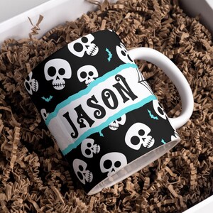 Può includere: Tazza in ceramica bianca con sfondo nero e un motivo di teschi bianchi. La tazza ha uno stendardo blu e bianco con il nome "Jason" scritto in nero.