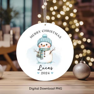 Könnte beinhalten: Ein weißer runder Weihnachtsbaumschmuck mit einem blauen Schneemann, der einen blauen Hut und einen Schal trägt. Der Schmuck trägt die Aufschrift "Frohe Weihnachten" und "Lucas 2024" in schwarzer Schrift.