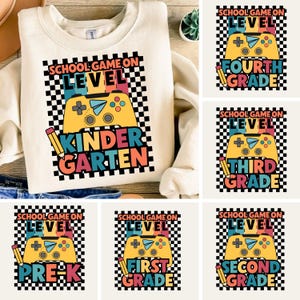 Puede incluir: Sudadera crema con un diseño gráfico colorido que presenta un mando de videojuegos y las palabras "School Game On Level Kindergarten". Imágenes adicionales muestran diseños similares para Pre-K a Cuarto Grado.
