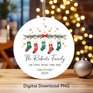May include: A white round Christmas bauble featuring a green and red garland of holly and berries with five red and green stockings hanging from it. The stockings have different patterns and designs. The text "The Roberts Family" is written in black script font below the stockings. The names "Liam, Sophie, Michael, Emma, Noah" are written in black below the family name. The year "2024" is written in black below the names.