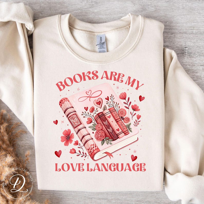 May include: A cream-coloured sweatshirt featuring the text "BOOKS ARE MY LOVE LANGUAGE" in red. The graphic shows a pink book with floral and heart details, and a stack of books. A comfortable design for book enthusiasts.