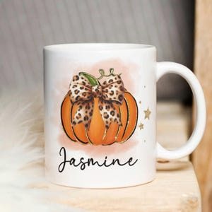 Può includere: Tazza in ceramica bianca con una zucca arancione con un fiocco leopardato. La zucca ha dettagli glitterati dorati ed è ambientata su uno sfondo acquerellato. Il nome "Jasmine" è scritto in corsivo.
