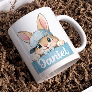May include: White ceramic mug with a cartoon bunny wearing a blue cap and bow tie. The bunny holds a blue sign with the name "Daniel" in a script font. The mug has a white handle and is set in a box with brown paper filler.