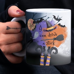 May include: White ceramic mug with a black and white design featuring a witch with a purple hat and a broom. The text on the mug reads "Yes! I can drive a stick".