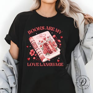 May include: Black t-shirt with the slogan "BOOKS ARE MY LOVE LANGUAGE" in red. The graphic shows a pink book with floral and heart details, and three stacked books. Ideal for book enthusiasts.