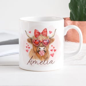 May include: White ceramic mug with a red and white bow, red heart-shaped sunglasses, and a red lollipop. The mug features a brown and white highland cow with the name "Amelia" printed below.