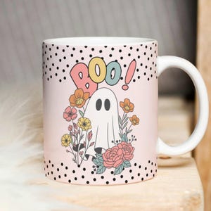 May include: A pink ceramic mug with a white ghost and floral design. The mug has black polka dots and the word "BOO!" in colourful letters.