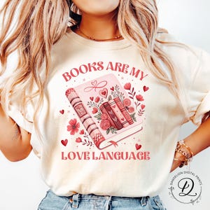 May include: Cream sweatshirt with the text "BOOKS ARE MY LOVE LANGUAGE" and a pink book illustration. The design includes floral and heart accents, and a stack of books. A romantic theme is present.