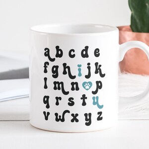 May include: White ceramic mug with a black alphabet design. The letter "i" is replaced with a blue paw print inside a heart. The text "I love" is written above the paw print.