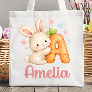 May include: White tote bag with a fluffy bunny and a carrot-shaped letter 'A'. The bag is decorated with the name 'Amelia' in pink script, and pink and blue flowers. A charming Easter-themed design.