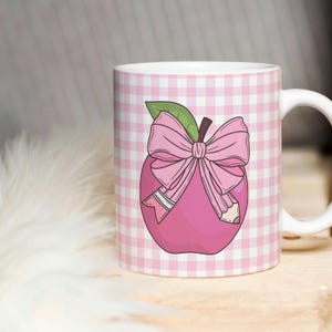 May include: Pink ceramic mug with a pink apple design. The apple has a pink bow and a pencil sticking out of it. The mug has a pink and white checkered pattern.