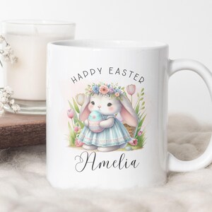 Puede incluir: Taza de cerámica blanca con una ilustración colorida de un conejito sosteniendo un huevo de Pascua. El texto "Happy Easter" está por encima del conejito y el nombre "Amelia" está por debajo del conejito.