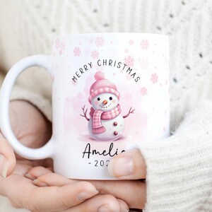 May include: White ceramic mug with a pink snowman design and the text "Merry Christmas". The snowman is wearing a pink hat and scarf. The mug also has the text "Amelia - 2022" on the bottom.