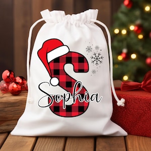 Puede incluir: Bolsa de regalo de Navidad blanca con una letra "S" de cuadros rojos y negros y el nombre "Sophia" en cursiva. La letra "S" tiene un gorro de Papá Noel rojo. Hay copos de nieve en la bolsa.