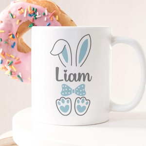 May include: White ceramic mug featuring a blue bunny design, a blue bow tie, and the name "Liam" printed on it.