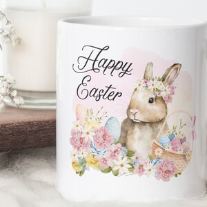 May include: White ceramic mug with a watercolor illustration of a bunny wearing a flower crown, a basket of Easter eggs, and the words "Happy Easter". The design features pastel pink, blue, yellow, and green floral elements.