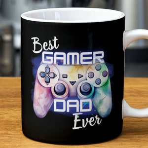 Puede incluir: Taza de café negra con asa blanca. La taza tiene una ilustración colorida de un mando de videojuego con el texto "Best Gamer Dad Ever".