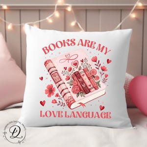 May include: White square cushion with the text "BOOKS ARE MY LOVE LANGUAGE" in red. The cushion features a pink book illustration with hearts, flowers, and other books. A decorative item for a bedroom or living room.