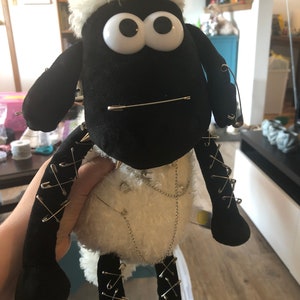 以下が含まれることがあります： 大きな白い目をした黒と白の羊のぬいぐるみ。おもちゃは安全ピンと銀色のチェーンで飾られています。羊は白い毛皮の襟をしています。