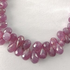 May include: A strand of pink gemstone beads, each bead is faceted and shaped like a teardrop. The beads are strung on a thin cord and are arranged in a loose, flowing pattern.