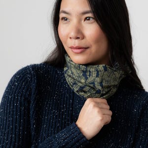 May include: A navy blue neck warmer featuring a fern design in olive green and navy. The snood is worn by a person wearing a navy blue jumper.