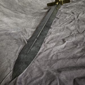 以下が含まれることがあります： 金色の柄と鍔を持つ、長い黒色の金属製の剣。剣は灰色の布の表面に置かれている。