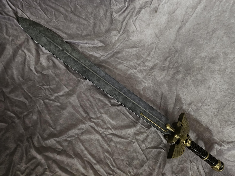 以下が含まれることがあります： 黒い柄と金色の鷲の装飾が施された銀色の剣。剣は灰色の布地に置かれている。