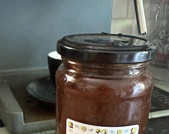 May include: A glass jar of apple butter with a black lid sits on a wooden cutting board. A white label with the words "Apple Butter" is affixed to the jar. The background includes a coffee machine.