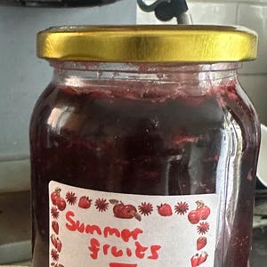 May include: A jar of dark red jam with a gold lid, labelled "Summer Fruits Jam." The label features red fruit illustrations. The jar is set against a blurred background, likely a kitchen.