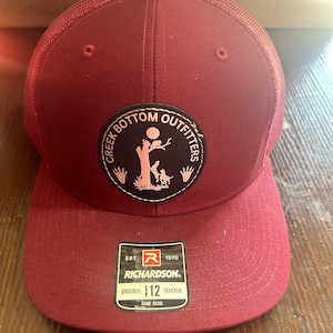 Op de afbeelding: Een bordeauxrode truckercap met een zwarte patch waarop in witte letters "Creek Bottom Outfitters" staat. De patch toont een roze silhouet van een boom met een vogel en twee mensen die eronder staan. De pet heeft een zwart label met de tekst "Richardson®" en "Original 112 Trucker" in witte letters.