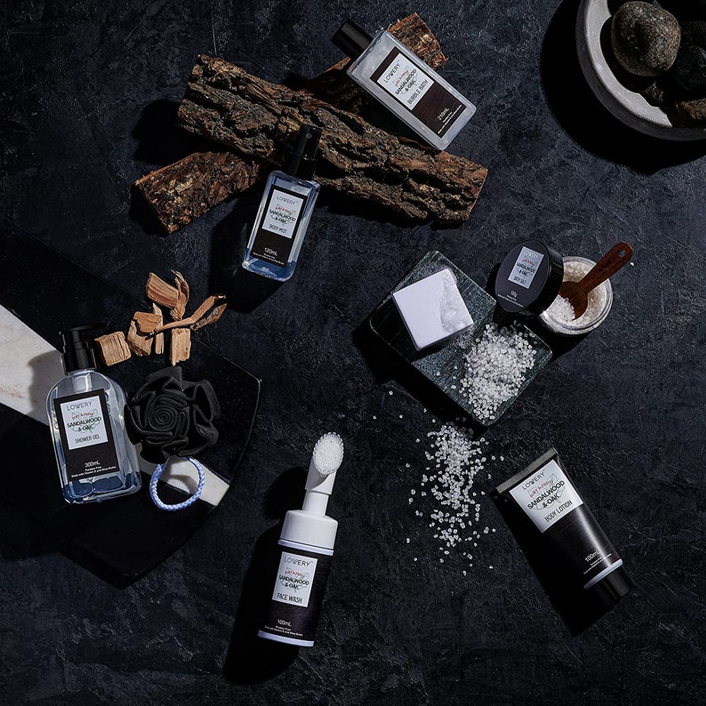 May include: An assortment of bath and body products, including shower gel, body mist, face wash, and body lotion, arranged on a dark surface. The products are labeled "LOVERY Sandalwood & Zinc". Other items include a black sponge, soap, and bath salts.