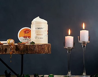 Può includere: Un barattolo bianco di burro corpo Lowerry al latte di mandorle e miele con un coperchio dorato. Il barattolo è appoggiato su una superficie di legno con un piccolo barattolo di miele e un mestolo di legno per il miele. Sullo sfondo ci sono due portacandele di vetro con candele bianche.
