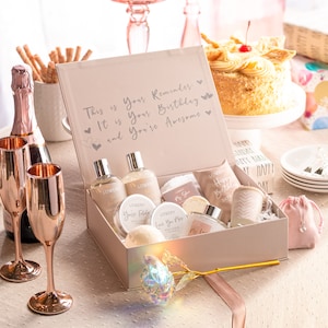 May include: A pink gift box filled with bath and body products, including body wash, lotion, bath bombs, and a bath towel. The box is decorated with a gold ribbon and a gold rose. The text on the box reads "This is your reminder: It is your birthday and you're awesome."