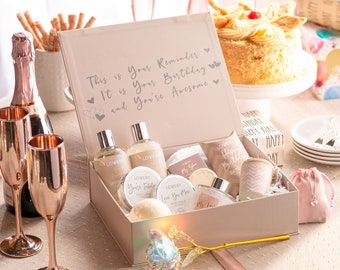 Può includere: Una scatola regalo rosa piena di prodotti per il bagno e il corpo, tra cui shampoo doccia, lozione, bombe da bagno e un asciugamano da bagno. La scatola è decorata con un nastro dorato e una rosa dorata. Il testo sulla scatola dice "Questo è il tuo promemoria: È il tuo compleanno e sei fantastico."