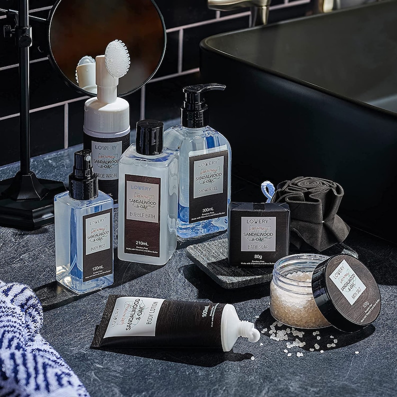 May include: A collection of sandalwood and oak-scented bath and body products, including body mist, bubble bath, shower gel, bath soap, body lotion, and bath salt. The products are in clear bottles and dark brown tubes with black labels. The products are arranged on a dark gray surface.