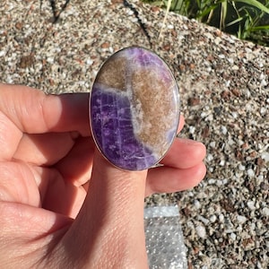 May include: Oval-shaped ring featuring a large, polished stone with a blend of purple and tan hues. The stone is set in a silver-colored metal band. The ring is held up by a hand, showcasing the ring's size and design.