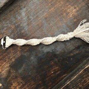 May include: A cream-colored macrame pacifier clip with a silver metal clip on one end and a looped tassel on the other. The twisted cord design is visible. The clip is designed to attach to a baby's clothing.