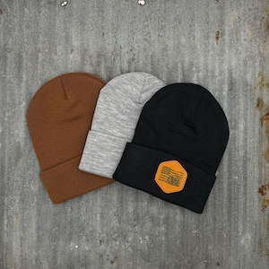 Op de afbeelding: Drie gebreide mutsen in bruin, grijs en zwart. De zwarte muts heeft een bruin leren patch met een Amerikaans vlagontwerp.