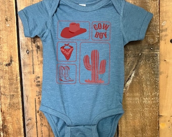 May include: A blue baby bodysuit with a red cowboy-themed graphic. The graphic includes a cowboy hat, the word "COWBOY", a bandana, cowboy boots, and a cactus. The bodysuit is hanging on a white hanger.