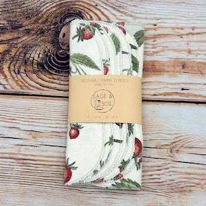 May include: A set of six reusable paper towels, each measuring 10 inches by 10 inches, with a strawberry and leaf pattern. The towels are made of 100% cotton and are wrapped with a brown paper band that reads "Sage & Soil".