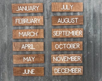 May include: Wooden rectangular plaques, each displaying a month of the year in white capital letters. The plaques are arranged in two columns against a textured, gray background. The wood has a natural, warm tone.