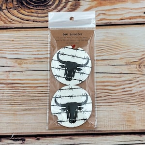 May include: Two car coasters with a black bull head design on a white background with barbed wire. The coasters are packaged in a clear plastic bag with a brown cardboard backing. Text on the backing reads "Car Coaster".